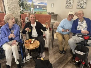 Four people, two elderly and two younger, sit together indoors, holding drinks and snacks, smiling at the camera during a cozy 3rd Anniversary event in a comfortable lounge setting.