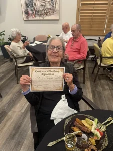 An elderly woman sitting at a dining table holds a Certificate of Residency Appreciation during the 3rd Anniversary Event. Other seniors are seated and eating in the background.