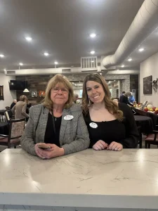 Two women wearing name tags stand side by side behind a marble counter in a restaurant or cafe, smiling at the camera during the 3rd Anniversary Event.