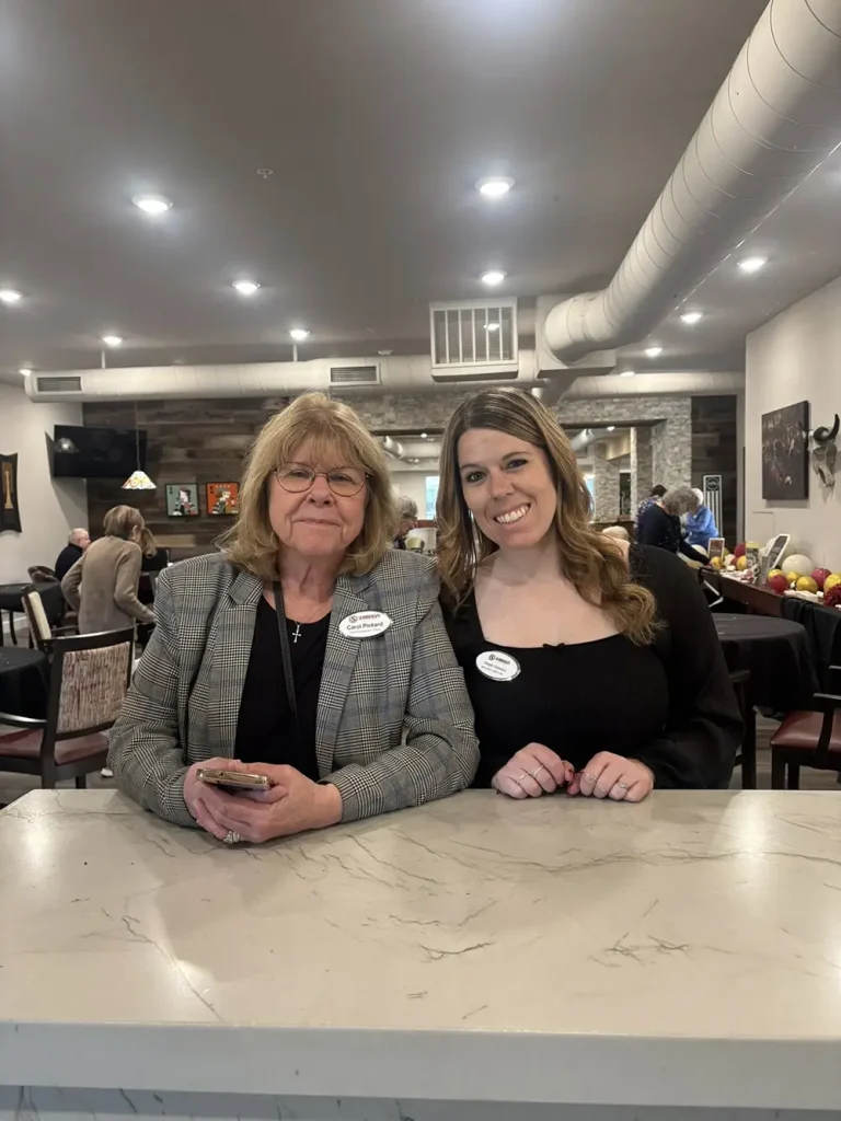 Two women wearing name tags stand side by side behind a marble counter in a restaurant or cafe, smiling at the camera during the 3rd Anniversary Event.