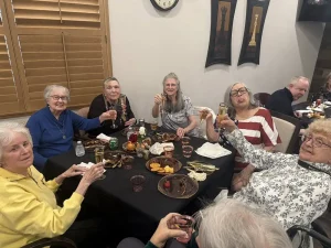 A group of older women sit around a dining table, holding up drinks and smiling as they celebrate a 3rd Anniversary Event, with finished plates and food items on the table.