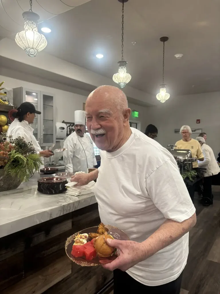 An older man in a white t-shirt holding a plate of food stands in a cafeteria buffet line during the 3rd Anniversary Event, with chefs and other people serving food in the background.
