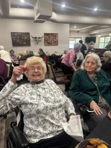 Two elderly women sit at a table in a crowded dining hall during the Anniversary Event; one smiles and holds up a small item, while others socialize in the background.