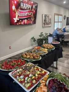 A buffet table with fruit, sliders, and appetizers is set up under a TV displaying "Happy Anniversary" at a 3rd Anniversary Event, with people seated in the background.
