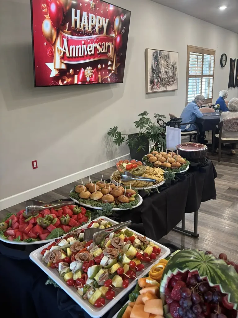 A buffet table with fruit, sliders, and appetizers is set up under a TV displaying "Happy Anniversary" at a 3rd Anniversary Event, with people seated in the background.