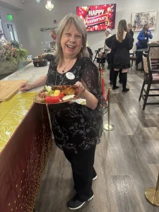 Smiling woman holds a plate of food at an indoor event with a "Happy Anniversary" sign in the background, celebrating a 3rd Anniversary as several people gather nearby.