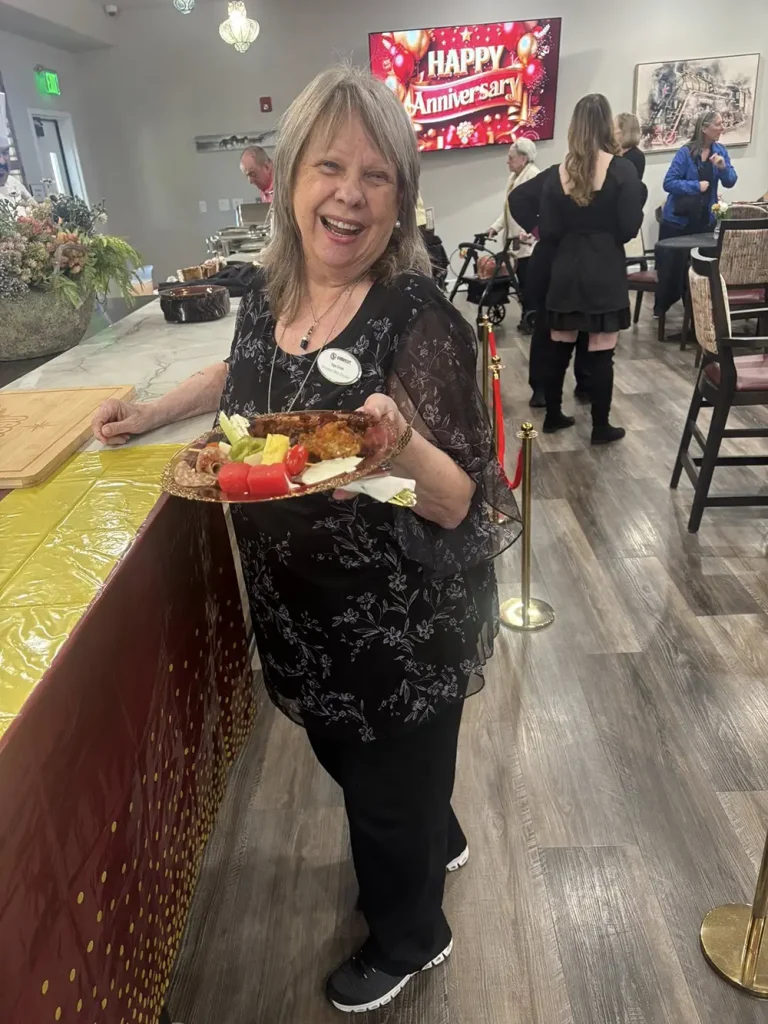Smiling woman holds a plate of food at an indoor event with a "Happy Anniversary" sign in the background, celebrating a 3rd Anniversary as several people gather nearby.