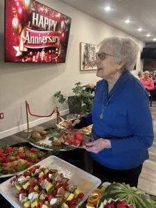 An elderly woman in a blue top serves herself fruit at the buffet table during an Anniversary Event, with a "Happy Anniversary" sign on a screen in the background.