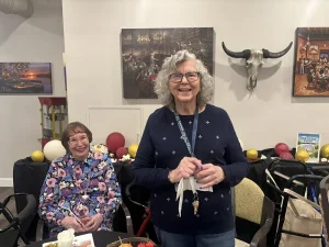 Two elderly women smile in a decorated room with balloons, a table of food, wall art, and a mounted cow skull, celebrating a joyful 3rd Anniversary event.