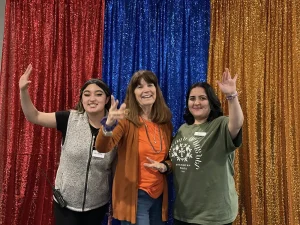 Three women stand and smile in front of glittery red, blue, and gold curtains—colors reflecting Armenian Heritage Month—each raising a hand with a peace sign or similar gesture.