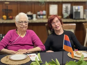 Two women sit at a dining table with plates of food and a small Armenian flag, celebrating Armenian Heritage Month in a warmly lit room with kitchen appliances in the background.