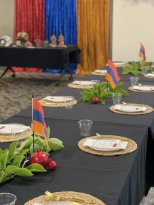 A table set with woven placemats, menus, greenery, red fruit, and small Armenian flags celebrates Armenian Heritage Month; colorful blue, red, and yellow curtains and a decorated table are in the background.