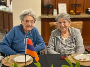 Two elderly women sit at a dining table with plates and drinks, smiling. An Armenian flag is displayed on the table in front of them, celebrating Armenian Heritage Month and the richness of Armenian culture.
