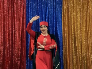 A woman in traditional red attire poses with one arm raised, standing in front of red, blue, and gold glittering curtains, celebrating Armenian Heritage Month.