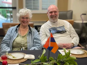 Two older adults sit side by side at a table set for a meal, with a small flag, plates, and greenery in front of them, celebrating Armenian Heritage Month.
