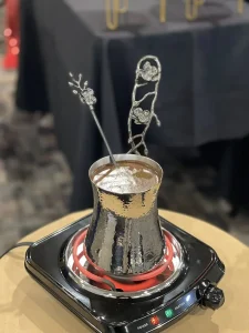 A metal Turkish coffee pot with ornate stirring sticks sits on an electric stovetop with the heating coil glowing red, evoking the warmth and tradition of Armenian heritage.