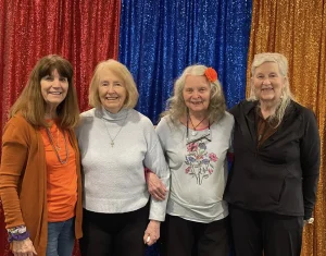 Four older women stand side by side, smiling at the camera in front of blue, red, and gold glittery curtains, celebrating Armenian Heritage Month and honoring Armenian culture.