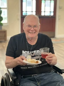 Elderly man in a wheelchair smiles while holding a plate of food and a cup of red drink. He wears a black Niemann-Pick Disease Awareness shirt, celebrating Armenian Heritage Month in a well-lit room.
