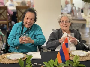 Two elderly women sit at a table holding drinks, smiling at the camera. A tricolor flag is on the table in front of them, with decorative greenery nearby, capturing a joyful moment during an Armenian Heritage Month celebration.