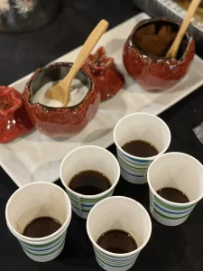 Five paper cups with dark liquid are arranged in front of two ceramic containers holding white and brown powders, each with a wooden spoon, set on a white tray—perfect for celebrating Armenian Heritage Month.