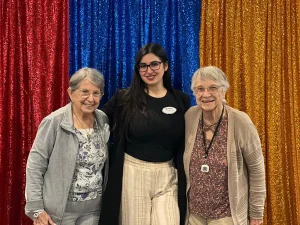 Three women pose and smile in front of red, blue, and yellow glittery curtains, celebrating Armenian Heritage Month. The woman in the center wears glasses and a name badge.