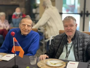 Two older men sit at a dining table with place settings, an Armenian flag between them, as they gather to celebrate Armenian Heritage Month. Other people are visible in the background.