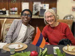 Two women sit at a table with place settings, smiling at the camera. In front of them are a small striped flag and a pomegranate, capturing a joyful moment during Armenian Heritage Month.
