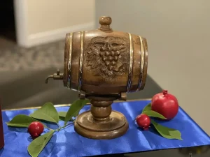 A small wooden barrel with a grapevine carving stands on a blue cloth, surrounded by green leaves and red pomegranates, beautifully celebrating Armenian heritage.