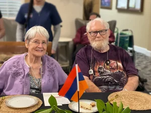 Two elderly people sit at a table with food and an Armenian flag, celebrating Armenian Heritage Month. Other people and plants are visible in the background.