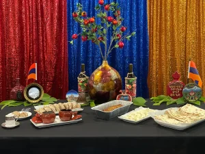 A table displays Armenian food and drinks, with a centerpiece fruit arrangement, Armenian flags, and a sequined red, blue, and yellow backdrop celebrating Armenian culture and history.