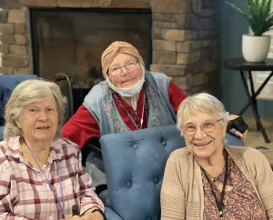 Three elderly women sit and smile indoors, celebrating Armenian Heritage Month, with a stone fireplace and a potted plant in the background. One woman wears a headscarf and a mask under her chin.