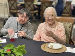 Two elderly women sit at a table, one smiling and holding a drink, the other pouring a red beverage. Plates, a placemat, and greenery are on the table as they celebrate Armenian Heritage Month together.