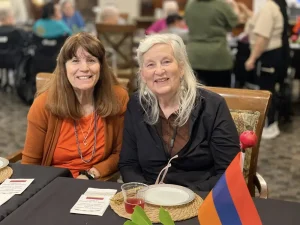 Two women are seated at a table with food and drinks, an Armenian flag displayed in front of them, celebrating Armenian Heritage Month. Other people are visible in the background.