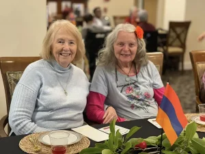 Two older women sit together at a decorated table with drinks and an Armenian flag, smiling at the camera in a communal indoor setting, celebrating Armenian Heritage Month and the richness of Armenian culture.