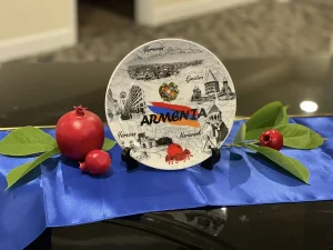 A decorative plate labeled "Armenia" with sketches of landmarks, placed on a stand, highlights Armenian culture. Pomegranates and green leaves rest on a blue cloth beneath the plate, celebrating rich tradition and artistry.