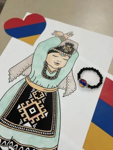 A drawing of a person in traditional Armenian clothing, a beaded bracelet with the Armenian flag, and paper hearts in the flag colors celebrate Armenian Heritage Month and honor Armenian culture on a table display.