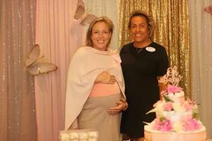 Two women stand in front of a sequin curtain with butterflies; one is pregnant. A pink and white "Oh Baby" cake with flowers sits on the table, setting the perfect scene for a joyful Baby Shower Celebration.