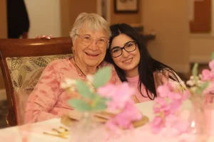 An elderly woman and a younger woman sit closely together at a decorated table, both smiling at the camera during a joyful baby shower celebration.
