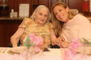 An elderly woman and a younger woman sit together at a table, smiling and holding hands, as they enjoy a Baby Shower Celebration surrounded by pink flowers and tableware in the foreground.
