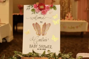 A decorative sign with flowers and butterflies reads "Welcome to Carlie's Baby Shower Celebration 04-30-2026" in a softly lit indoor setting.