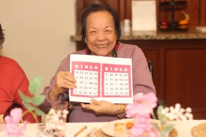 An elderly woman smiles while holding up two bingo cards at a table decorated with flowers and food during a Baby Shower Celebration.