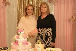Two women stand in front of a gold and pink backdrop at a decorated table with cakes and flowers—one pregnant and smiling, the other beside her—capturing a joyful Baby Shower Celebration.