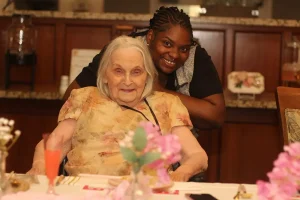 An elderly woman sits at a table with flowers, smiling, while a younger woman stands behind her, also smiling, in a warmly lit room during a joyful Baby Shower celebration.