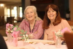 Two older women sit side by side at a table decorated with pink flowers and vases, smiling at the camera in a warmly lit room during a joyful Baby Shower Celebration.