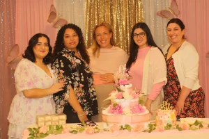Five women stand together behind a decorated table at a Baby Shower Celebration, with a cake and pink floral decorations, smiling at the camera.
