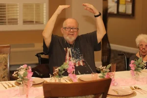 An older man sits at a decorated dining table with pink flowers, raising his arms above his head, as an elderly woman nearby looks on during a joyful Baby Shower Celebration.