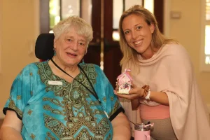 An older woman in a wheelchair sits next to a smiling younger woman holding a small, decorative pink house at a joyful baby shower celebration.