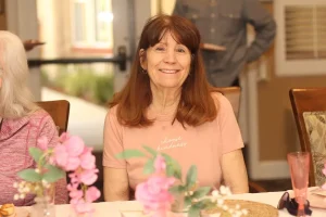 A woman with long brown hair sits at a table decorated with pink flowers, smiling at the camera during a baby shower celebration. She is wearing a light pink shirt that reads "choose kindness.