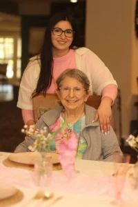 A young woman stands behind an elderly woman seated at a decorated table indoors, both smiling at the camera during a joyful Baby Shower Celebration.