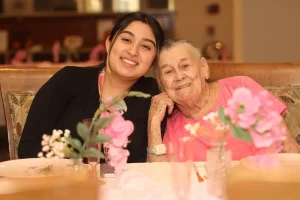 A young woman and an elderly woman sit together at a table decorated with pink flowers, smiling at the camera during a joyful baby shower celebration.